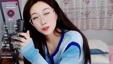 Media: A video of an Asian woman with long, straight black hair, wearing glasses and a blue sweater, holding a smartphone in a bedroom with a pink and white floral-patterned curtain.