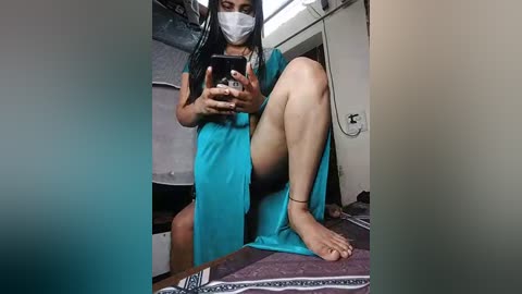 Media: Video of a woman in a teal towel, wearing a white face mask and black shorts, sitting on a bed with a patterned blanket.