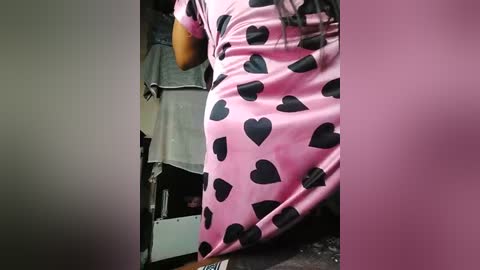 Media: A video of a person wearing a pink dress with black heart patterns, standing in a cluttered room with a grey towel and various objects on the floor.