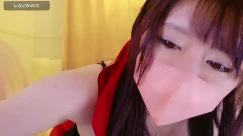 Media: Video of a young Asian woman with straight black hair and fair skin, wearing a red top, blowing a kiss. Background is a yellow curtain, possibly in a bedroom setting.