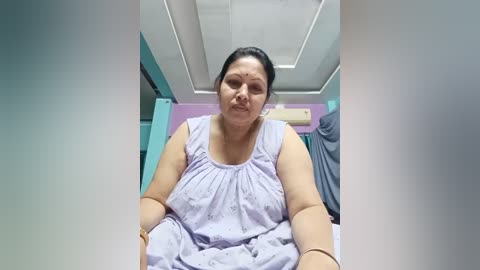 Media: Video of a middle-aged woman with medium skin tone, dark hair tied back, wearing a light purple blouse, sitting in a medical exam room with teal and white walls and an air conditioner.