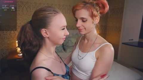 Media: Video of two young women with fair skin, one in a black tank top, the other in a white crop top, in a cozy bedroom with a tiled wall and lit lamp.