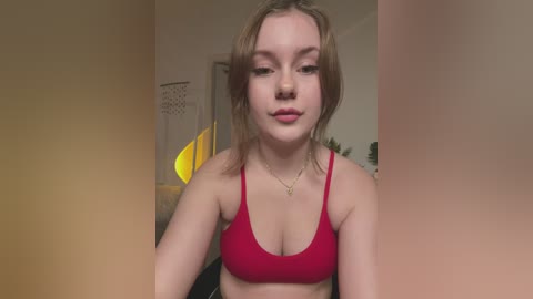 Media: Video of a young woman with fair skin and shoulder-length brown hair, wearing a red sports bra and a gold necklace, standing indoors against a beige wall.