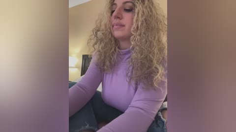 Media: Video of a fair-skinned woman with curly blonde hair, wearing a tight lavender long-sleeve top, sitting on a couch in a dimly lit room.