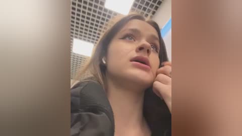 Media: A video of a young Caucasian woman with light skin and straight brown hair, wearing a black top, leaning against a beige wall in a cubicle with a grid-patterned ceiling.