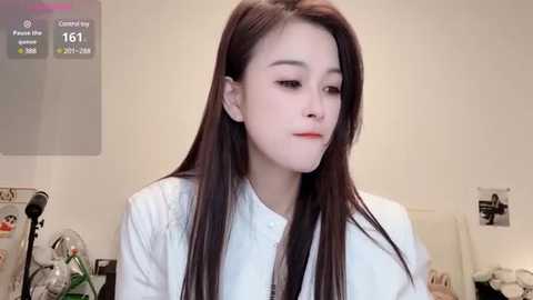 Media: A video of a young Asian woman with long, straight black hair, pale skin, and light makeup, wearing a white blouse, in a minimalistic room with a calendar and plants.