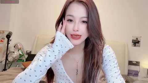 Media: Video of an Asian woman with long, straight brown hair, wearing a white polka-dot top, talking on a phone, in a cozy bedroom with a beige headboard and white walls.