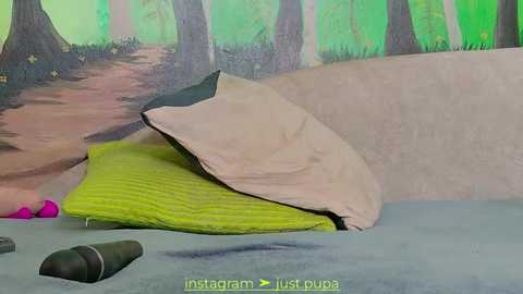 Media: Video of a cozy bedroom scene with a beige pillow on a green bedspread. Background features a painted forest mural. A black microphone and a pink object are visible in the foreground.