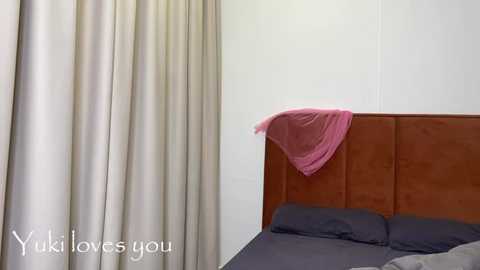 Media: Video of a minimalist bedroom with beige curtains, a brown upholstered headboard, and a bed covered in dark grey sheets. A pink, draped garment hangs from the headboard. Text reads \"Yukiloves you\" in the bottom left corner.