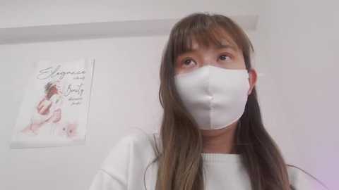 Media: Video of an East Asian woman with long brown hair, wearing a white surgical mask, white top, standing in a sterile white room with a framed art poster of a baby on the wall.