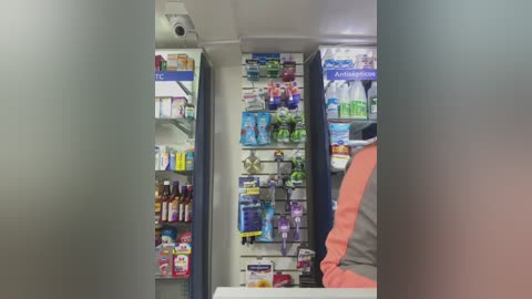 Media: Video of a modern pharmacy interior with an aisle of neatly organized medicine shelves and a person in a white coat and orange apron, likely a pharmacist, standing in the background.