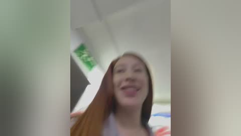 Media: A video of a young woman with long, straight brown hair, smiling widely, taken from a low angle, showing a blurry background with white walls and a green sign.