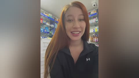 Media: Video of a smiling Asian woman with long red hair, wearing a black Under Armour hoodie, standing in a brightly lit pharmacy with shelves filled with various products.