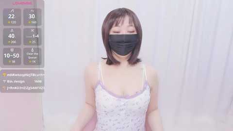 Media: A video of a young Asian woman with short brown hair, wearing a black face mask, white spaghetti-strap dress, and light pink panties. She stands against a white curtain background, with a digital overlay showing \"Live\" and heart icons.