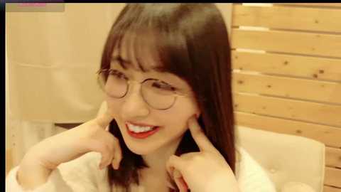 Media: Video of an Asian woman with straight black hair, wearing glasses, red lipstick, and a white shirt, smiling and touching her ears, in a cozy, wooden-paneled room.