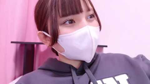 Media: Video of an Asian woman with straight brown hair, wearing a white surgical mask, gray hoodie, and sitting against a pink wall.