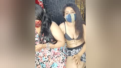 Media: Video of two women in a bedroom; one with long black hair, wearing a floral dress, and a face mask; the other, with long dark hair, wearing a black bra and plaid shorts.