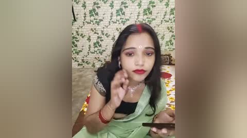 Media: Video of a young South Asian woman in traditional attire, green sari, black blouse, red bindi, red lipstick, sitting on a floral bedspread, against a green ivy-covered brick wall.