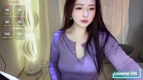 Media: A video of an Asian woman with long dark hair, fair skin, and large breasts, wearing a lavender long-sleeve shirt, sitting in a modern room with a lit lamp and beige curtains.