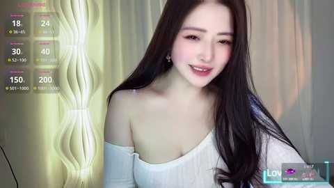 Media: Video of a young East Asian woman with long black hair, fair skin, and a slender physique, wearing a white off-shoulder top, smiling. Background features a soft, glowing light and a live stream overlay.