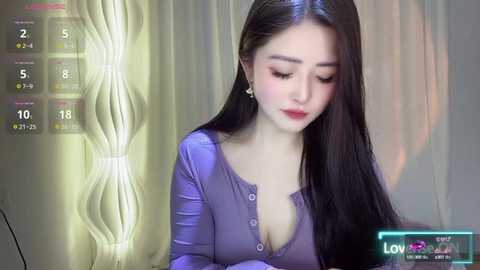 Media: A video of a young East Asian woman with long black hair, wearing a purple long-sleeve top with a deep neckline, standing in a dimly lit room with a wavy, light-colored wall.