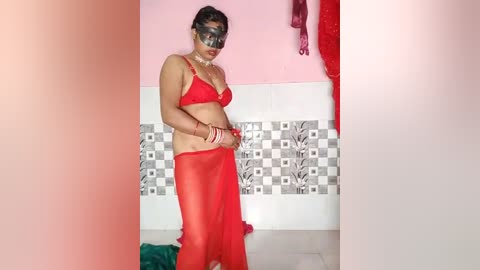 Media: Video of a South Asian woman in a red saree, wearing a red bra, and black sunglasses, standing in a pink bathroom with white and gray tile accents.