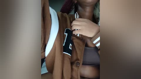 Media: Video of a woman with light brown skin, wearing a brown jacket over a white sports bra, with a black football number \"1\" visible. Her left hand is on her chest, and her lips are partially visible.