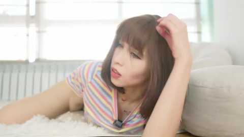 Media: Video of a young woman with light skin, straight brown hair, and a striped shirt, lying on a white rug in a bright, minimalist room with large windows.