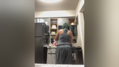 Media: Video of a woman with braided hair in a gray tank top and patterned pants, preparing food in a modern, well-lit kitchen with white walls, black appliances, and neatly arranged shelves.