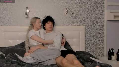 Media: Video of a couple in a modern, patterned bedroom; a blonde woman in a white dress, a dark-haired man in a white shirt, cuddling on a grey bedspread.