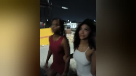 Media: A blurred video of two young women walking at night, one in a red dress, the other in a white tank top. Background includes a yellow barrier and dimly lit buildings.