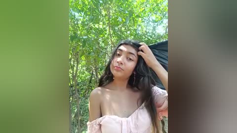 Media: Video of a young woman with long black hair, light skin, and medium build, wearing a pink off-shoulder dress, standing in a lush, green forest, holding a black umbrella.