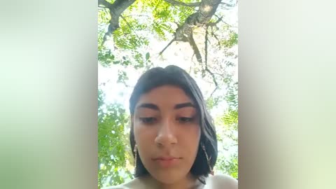 Media: Video of a young woman with medium brown skin, shoulder-length black hair, and thick eyebrows, wearing a white top. Background features bright green foliage and sunlight filtering through the leaves.