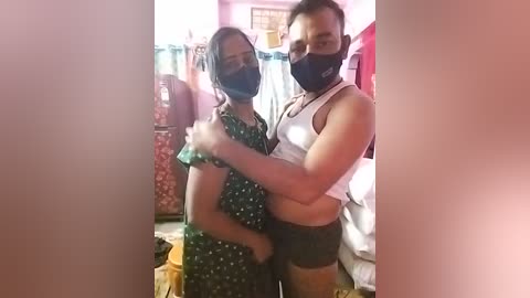 Media: Video of two people, one in a black mask and green dress, the other in a white tank top and black shorts, embracing in a cluttered room with pink walls and various items.