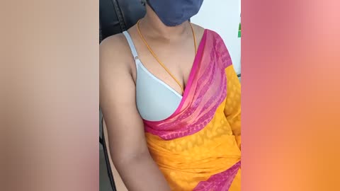 Media: Video of a woman with medium-brown skin, wearing a light blue bra, yellow-orange sari, and blue mask, seated indoors.