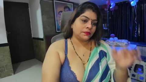 Media: Video of a South Asian woman with medium skin tone and dark hair, wearing a blue and white striped sari with a low-cut neckline, holding a blue cake in a dimly lit room with framed photos and balloons.