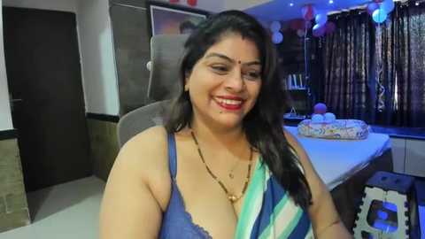 Media: A video of a smiling South Asian woman with medium skin tone, dark hair, wearing a blue and green saree, seated indoors at a birthday party.