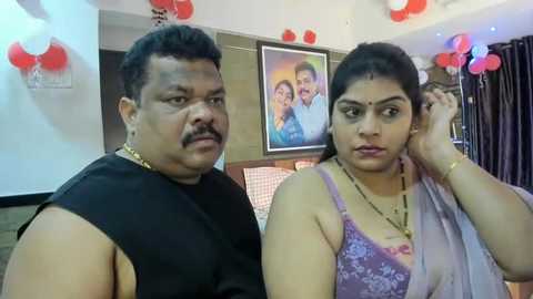 Media: Video of an Indian couple in casual attire, the man in a black sleeveless shirt and the woman in a purple floral blouse. They are indoors, with red balloons, a framed family photo, and a checkered table visible in the background.