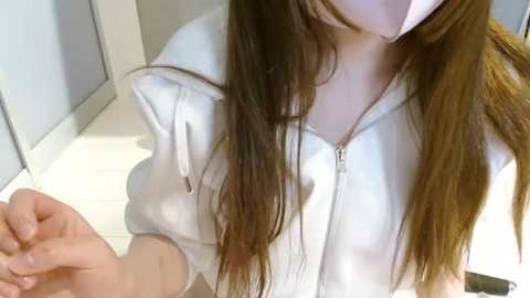 Media: Video of a woman with long brown hair, wearing a white hoodie, kneeling on a white tiled floor. She holds a black object in her right hand.