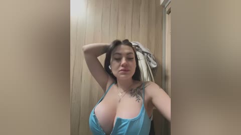 Media: Video of a light-skinned woman with long brown hair, wearing a low-cut blue tank top, revealing large breasts, and a tattoo on her left shoulder. She stands in a wooden-paneled room with beige walls.