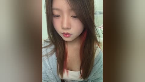 Media: A video of a young Asian woman with long brown hair, red highlights, and pale skin, wearing a white top and light blue cardigan, looking down with a pensive expression.