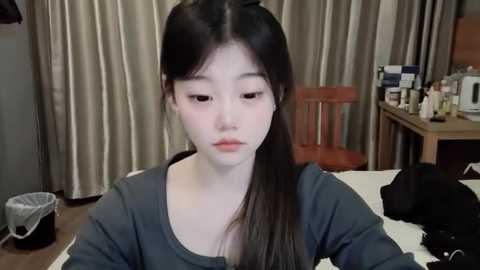 Media: A video of an Asian woman with pale skin and long black hair, sitting at a desk in a cluttered room. She wears a gray shirt and looks downcast.