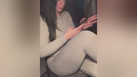 Media: A video captures a young woman with long, straight black hair, wearing a beige top and tight, light-gray leggings, seated on a chair, holding a smartphone. The background is blurry.