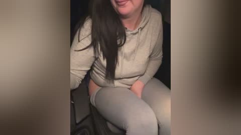 Media: A video of a middle-aged woman with long dark hair, wearing a beige hoodie and gray leggings, sitting in a dark room with blurry background.