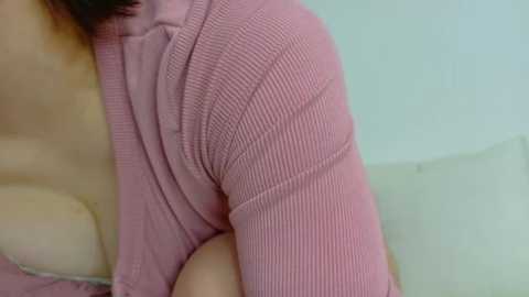 Media: Video of a light-skinned woman's upper chest and shoulder, wearing a pink, ribbed, cropped sweater, revealing her medium-sized breasts. Background is a light-colored wall.