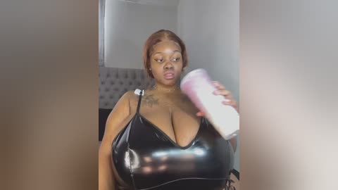 Media: Video of a plus-size Black woman with light brown skin, wearing a black latex dress with large cleavage, holding a pink dildo, in a modern, minimalist room with a tufted headboard.