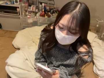 Media: Video of an Asian woman with long dark hair, wearing a white face mask, gray sweater, and holding a smartphone, seated on a beige couch in a cluttered living room.