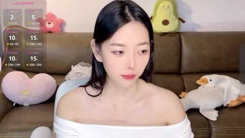 Media: Video of an East Asian woman with fair skin, black hair, and red lipstick, sitting on a brown leather couch. Background includes plush toys, a calendar, and a green dinosaur.