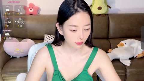 Media: Video of an Asian woman with straight black hair, wearing a green sleeveless top, sitting on a brown couch. Background includes stuffed toys, a virtual game screen, and a plush pillow.