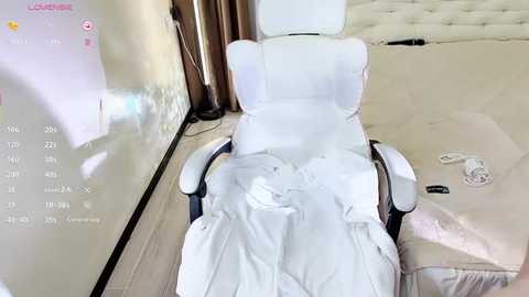 Media: A video of a white gaming chair covered in white sheets, positioned in a modern room with beige walls and a tufted leather sofa, with a digital clock on the wall displaying the time.